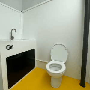 Intérieur d’un Bio’loo : cabine sanitaire équipée d’une toilette à cuvette blanche et d’un lave-mains intégré à structure blanche, installés sur un sol jaune vif.
